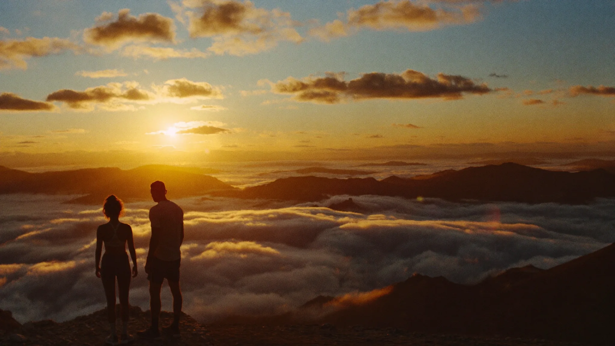 Eine Frau und ein Mann in Sportkleidung stehen auf einem Gipfel über dem Wolkenmeer und schauen in den Sonnenuntergang.